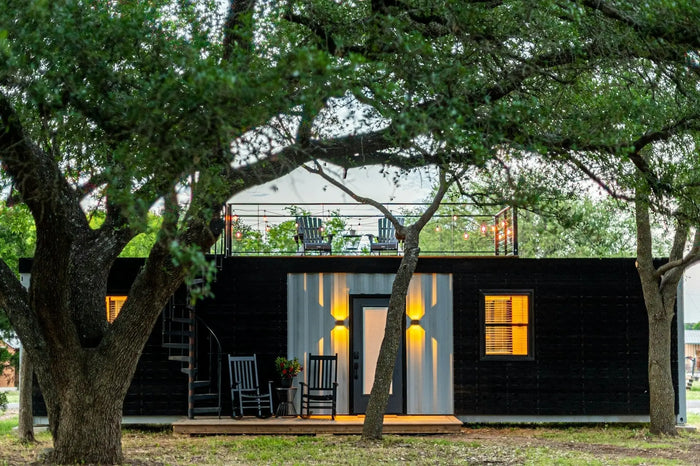 Black and white corrugated metal shipping container home with balcony porch chairs and warm interior lighting