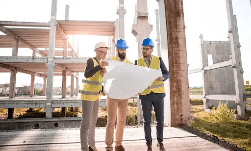Three construction workers in blue hard hats review a detailed site plan near a concrete structure for building permit decisions.