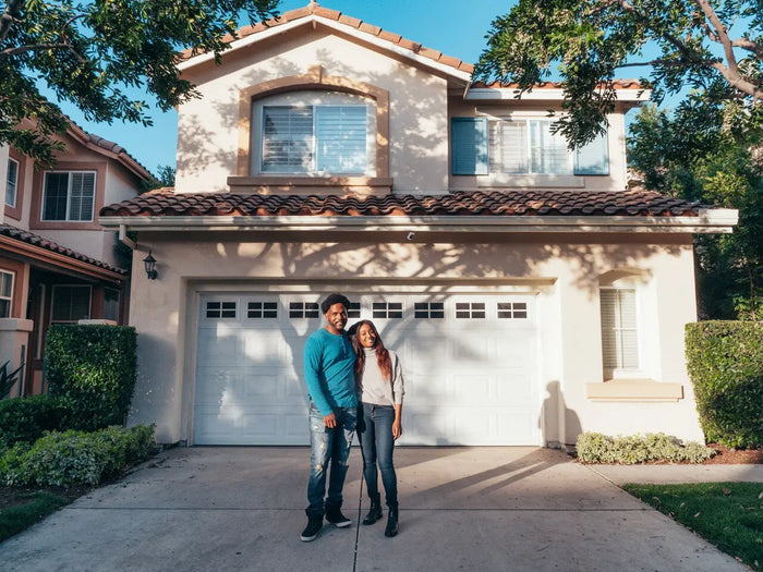 A couple stands in front of a beige stucco house with red tile roofing featuring a second story addition.