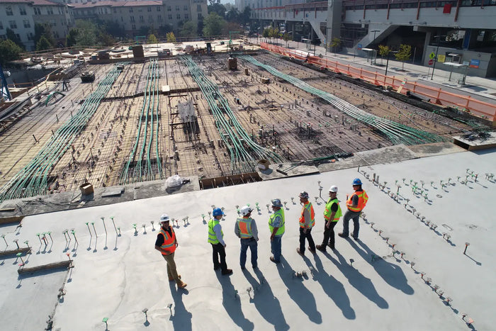 Six construction workers in high-visibility vests on concrete slab reviewing site plan for design-bid-build process projects.