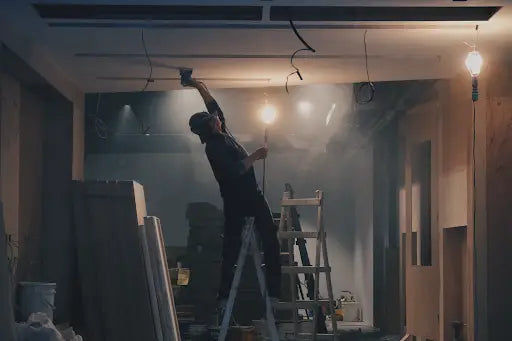 A worker in dark clothing installs wiring on a ladder in an unfinished room while referencing a site plan for load-bearing walls.