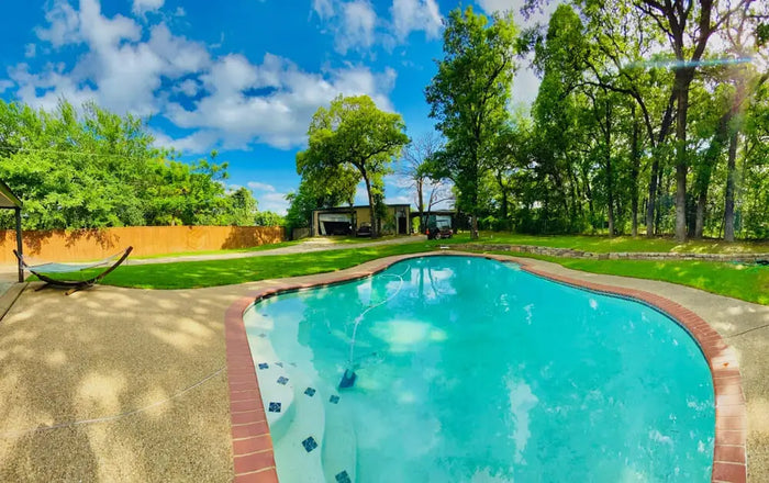 A clear turquoise inground pool with reddish brown tile edging surrounded by grass and trees illustrating pool costs.