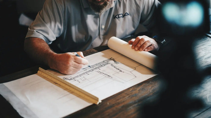 A man in a gray polo shirt drawing architectural drawings with a blue pen and ruler at a wooden table.